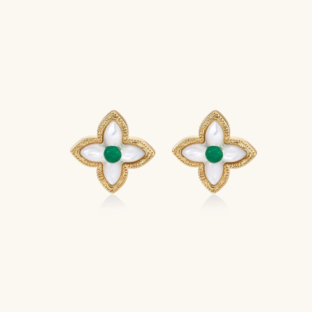 Gold quatrefoil stud earrings with white mother-of-pearl petals and emerald green gemstone center on beige background.