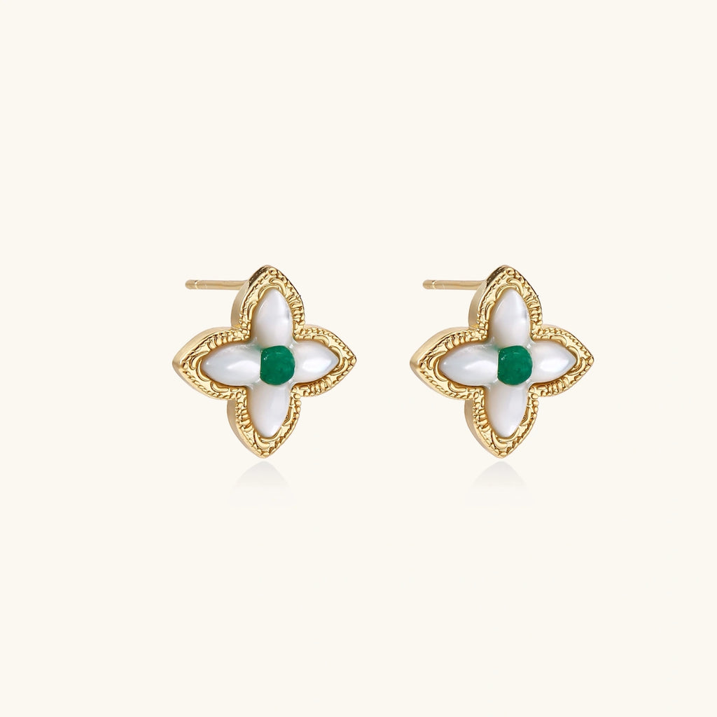 Gold quatrefoil stud earrings with white mother-of-pearl petals and emerald green gemstone center on beige background.